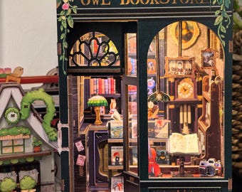 Może przedstawiać: Szczegółowy miniaturowy model Owl Bookstore. Ciemnozielona fasada z napisem "OWL BOOKSTORE" i kwiatowymi dekoracjami. Wewnątrz miniaturowe książki wypełniają półki, obok zegara i lampy, tworząc ciepłą, zachęcającą atmosferę. Model ma uroczy, szczegółowy design.