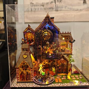 May include: A detailed miniature dollhouse with multiple levels, featuring intricate architectural details and warm interior lighting. The facade includes a clock tower, arched windows, and decorative elements. The scene is enclosed in a clear display case.