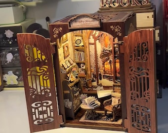 Może przedstawiać: Miniaturowy drewniany model księgarni z otwartymi drzwiami, prezentujący szczegółowe wnętrze z półkami na książki, stołem i krzesłem. Model posiada skomplikowane wycinane wzory, a na górze napis „BOOKS”. Wnętrze jest oświetlone, tworząc ciepłą, zachęcającą scenę.