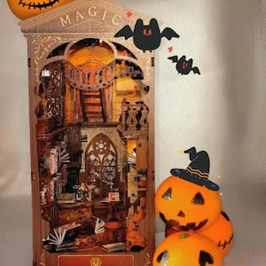 May include: A wooden Magic Book House with interior details, lit with warm light. The house is decorated with a jack-o'-lantern, bats, and pumpkins wearing a witch hat, creating a Halloween theme. The text "MAGIC" and "MAGIC BOOK HOUSE" are visible.