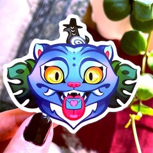 Può includere: Un adesivo raffigurante un gatto blu con grandi occhi gialli, un naso rosa e una lingua con un disegno a forma di cuore. Il gatto ha un cappello nero e dettagli di foglie verdi. L'adesivo è tenuto da una mano con smalto nero.