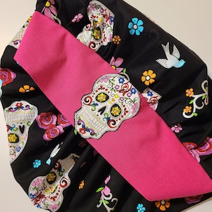 May include: A black fabric scrunchie with a pink ribbon and a colorful sugar skull pattern. The fabric features a repeating pattern of sugar skulls, flowers, and birds.