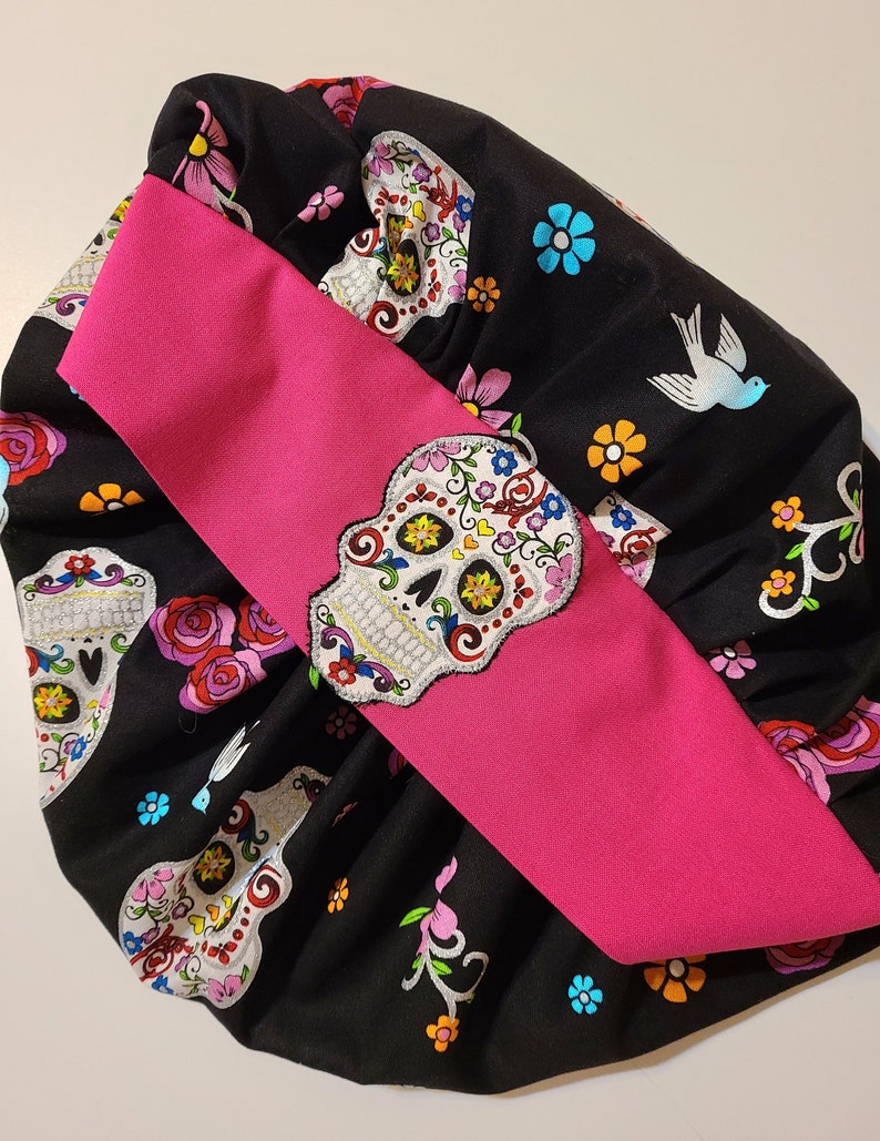 May include: A black fabric scrunchie with a pink ribbon and a colorful sugar skull pattern. The fabric features a repeating pattern of sugar skulls, flowers, and birds.