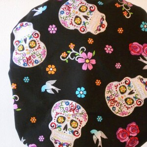 May include: Black fabric with a repeating pattern of colorful sugar skulls and flowers. The skulls have white eyes and are decorated with floral designs. There are also small blue birds scattered throughout the fabric.