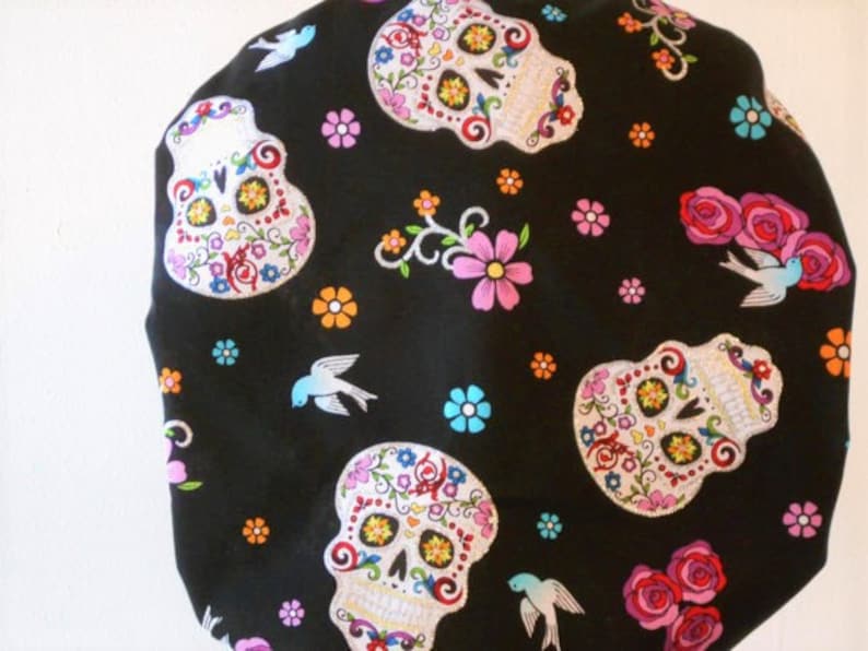 May include: Black fabric with a repeating pattern of colorful sugar skulls and flowers. The skulls have white eyes and are decorated with floral designs. There are also small blue birds scattered throughout the fabric.