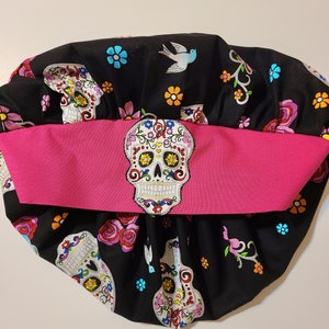 May include: Black fabric scrub hat with a pink band and a colorful sugar skull design. The hat has a gathered top and is decorated with flowers and birds.