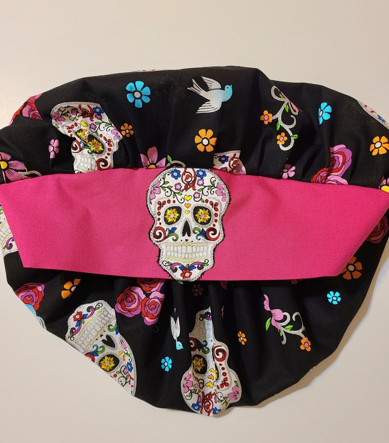 May include: Black fabric scrub hat with a pink band and a colorful sugar skull design. The hat has a gathered top and is decorated with flowers and birds.