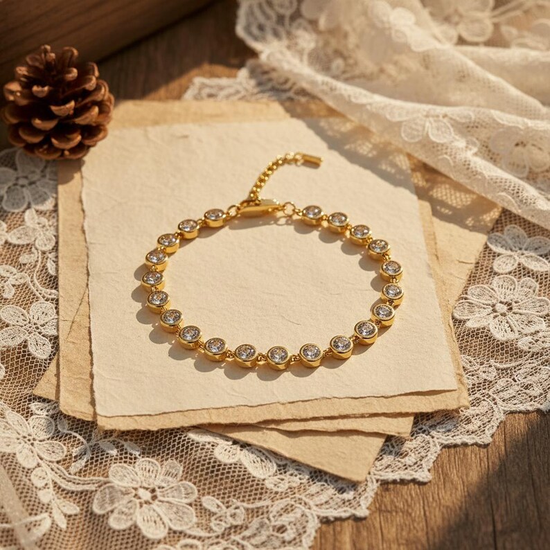 May include: A gold-toned bracelet with clear, round gemstones set in a bezel design. The bracelet is displayed on a stack of textured paper, with a pinecone and lace in the background. The jewelry is a delicate accessory.
