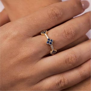 May include: A gold ring with a repeating pattern of four-leaf clover designs. Each clover is made of dark blue gemstones, with small white gemstones between the clovers. The ring is on a person's finger.