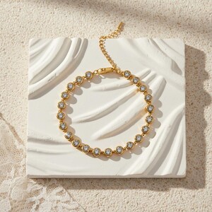 May include: A gold-colored bracelet with a series of clear, round gemstones set in individual bezels. The bracelet is displayed on a white, textured surface, showcasing its delicate design and sparkle. The clasp and chain are also gold.