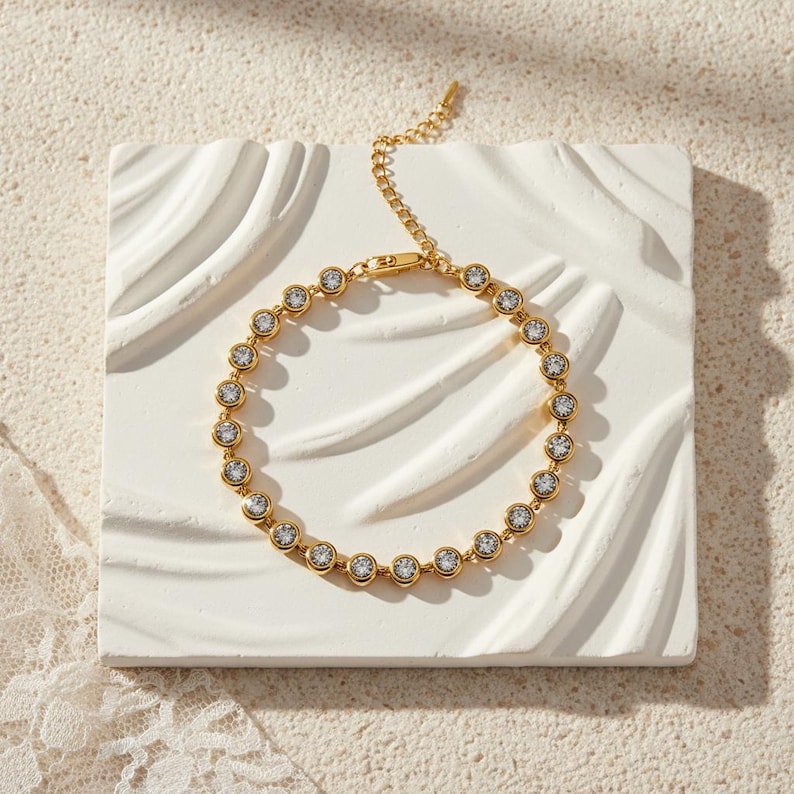 May include: A gold-colored bracelet with a series of clear, round gemstones set in individual bezels. The bracelet is displayed on a white, textured surface, showcasing its delicate design and sparkle. The clasp and chain are also gold.