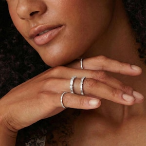 Puede incluir: Primer plano de una mano que muestra varios anillos de plata. Los anillos presentan varios diseños, incluyendo bandas con piedras pequeñas y espaciadas uniformemente y anillos con piedras rectangulares más grandes. El tono de piel es medio, con un fondo borroso.