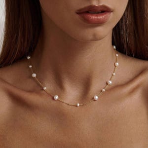 Op de afbeelding: Fijne gouden ketting met een reeks witte parels. De parels variëren in grootte en zijn geregen aan een fijne gouden ketting. De ketting wordt getoond om de nek van een persoon, wat het elegante ontwerp benadrukt.