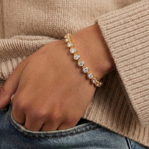 May include: A gold-toned bracelet with clear, round gemstones. The bracelet is worn on a wrist, with a neutral-colored sweater and denim jeans in the background. The gemstones are evenly spaced along the bracelet.