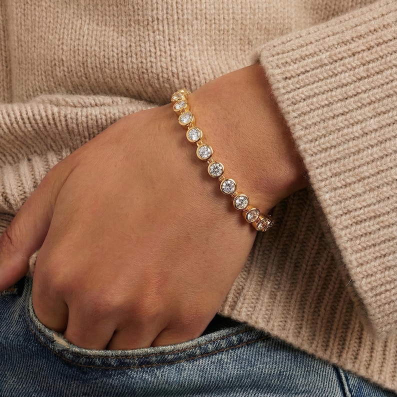 May include: A gold-toned bracelet with clear, round gemstones. The bracelet is worn on a wrist, with a neutral-colored sweater and denim jeans in the background. The gemstones are evenly spaced along the bracelet.