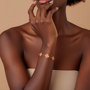 May include: A gold bracelet featuring a series of square-shaped charms with a pink, textured design. The bracelet is worn on the arm of a person with dark skin, against a neutral background. The bracelet's design is intricate and delicate.