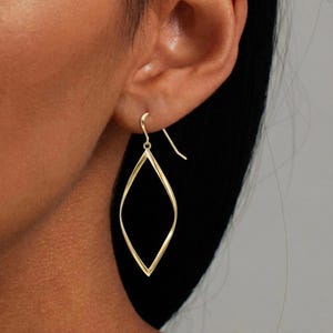 Könnte beinhalten: Ein Paar goldfarbene Ohrringe mit offenem Rahmen. Jeder Ohrring hat eine Tropfenform mit einem schlanken, gedrehten Design. Die Ohrringe hängen an einem Haken, der durch das Ohr geht.