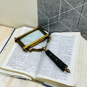 Pode incluir: Um livro aberto com páginas envelhecidas, com uma lupa com moldura de latão e uma lupa com uma alça preta esculpida. O livro está sobre uma superfície clara, com um fundo com padrão geométrico.