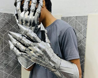 Puede incluir: Guanteletes de metal plateado con largas extensiones puntiagudas para los dedos. Los guanteletes tienen grabados detallados y se llevan en los brazos. La persona de la imagen lleva una camiseta gris.