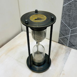 Puede incluir: Un reloj de arena de latón antiguo con un centro de vidrio, lleno de arena. El reloj de arena está sostenido por un marco y una base de metal oscuro. Una placa circular de latón en la parte superior tiene texto y una ilustración de un barco. El reloj de arena está sobre una superficie de madera clara.