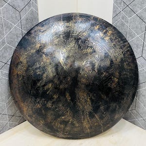 May include: A large, round, metal decorative plate with an intricate, embossed pattern. The plate has a dark, aged finish with gold accents, creating a vintage aesthetic. The design is symmetrical and detailed, suitable for wall decor.