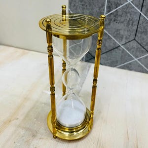 May include: A brass hourglass with a clear glass center filled with white sand. The hourglass has a circular base and top, with decorative vertical supports. The design is vintage and elegant.