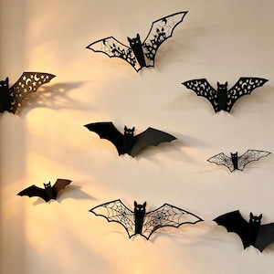 May include: A collection of black bat-shaped wall decorations in various sizes. Some bats feature intricate cut-out designs including spider webs, crescent moons, stars, and silhouettes of haunted houses, while others are solid black shapes.