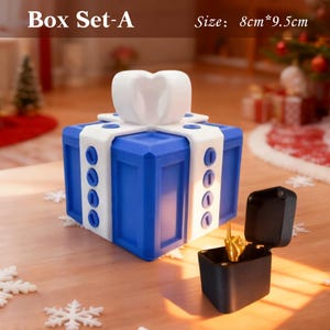 May include: A blue gift box with white ribbon accents and a white bow on top. The box is 8 cm x 9.5 cm. A small black box with a gold-colored hand gesture is open next to the gift box. The text "Box Set-A" is visible.