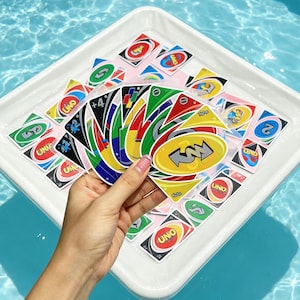 Pode incluir: Uma mão segurando um leque de cartas de jogo Uno coloridas com vários números e símbolos de ação, incluindo uma carta de mais quatro. Cartas Uno adicionais estão espalhadas por uma bandeja branca, com designs vermelhos, azuis, verdes e amarelos.
