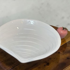 May include: A white ceramic bowl shaped like a large clam shell with concentric ridges along the interior surface. Beside the bowl sits a small, glossy pink decorative apple with a brown stem, both resting on a polished dark wood surface.