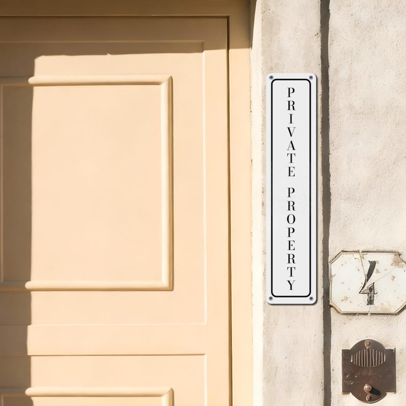 May include: A rectangular white metal sign with the words "PRIVATE PROPERTY" in black, mounted on a textured wall. The sign is vertically oriented and framed with a thin black border. A cream-colored door is visible in the background.