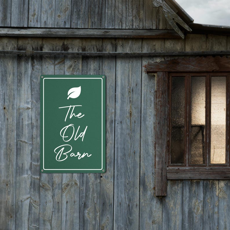 May include: A rectangular green metal sign with white text that reads "The Old Barn" is mounted on a weathered wooden wall. A white leaf graphic is above the text. A window is visible to the right.