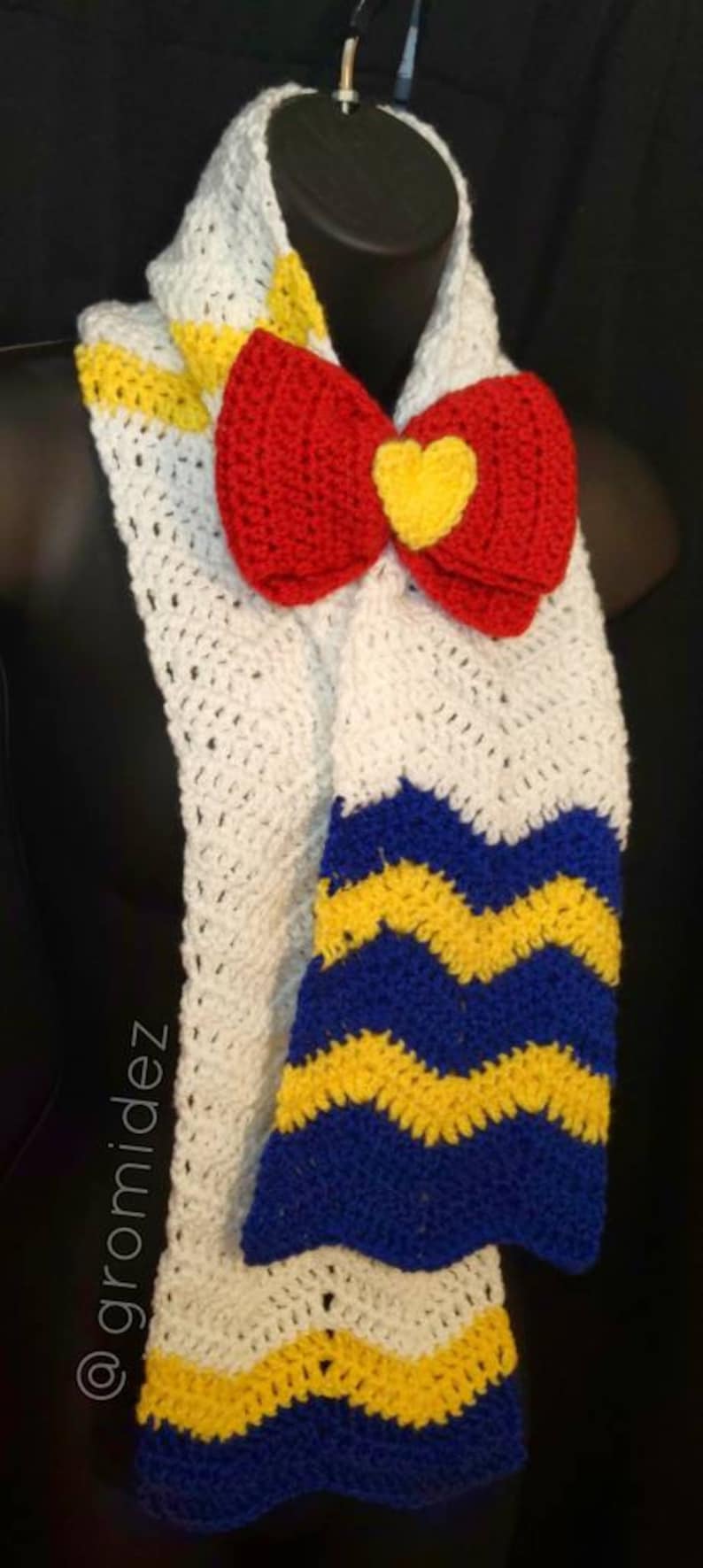 May include: White crocheted scarf with a red and yellow bow and blue and yellow chevron stripes.