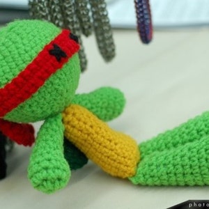 May include: Green crocheted turtle with a red and black mask. The turtle is lying on its side with its legs and tail outstretched.