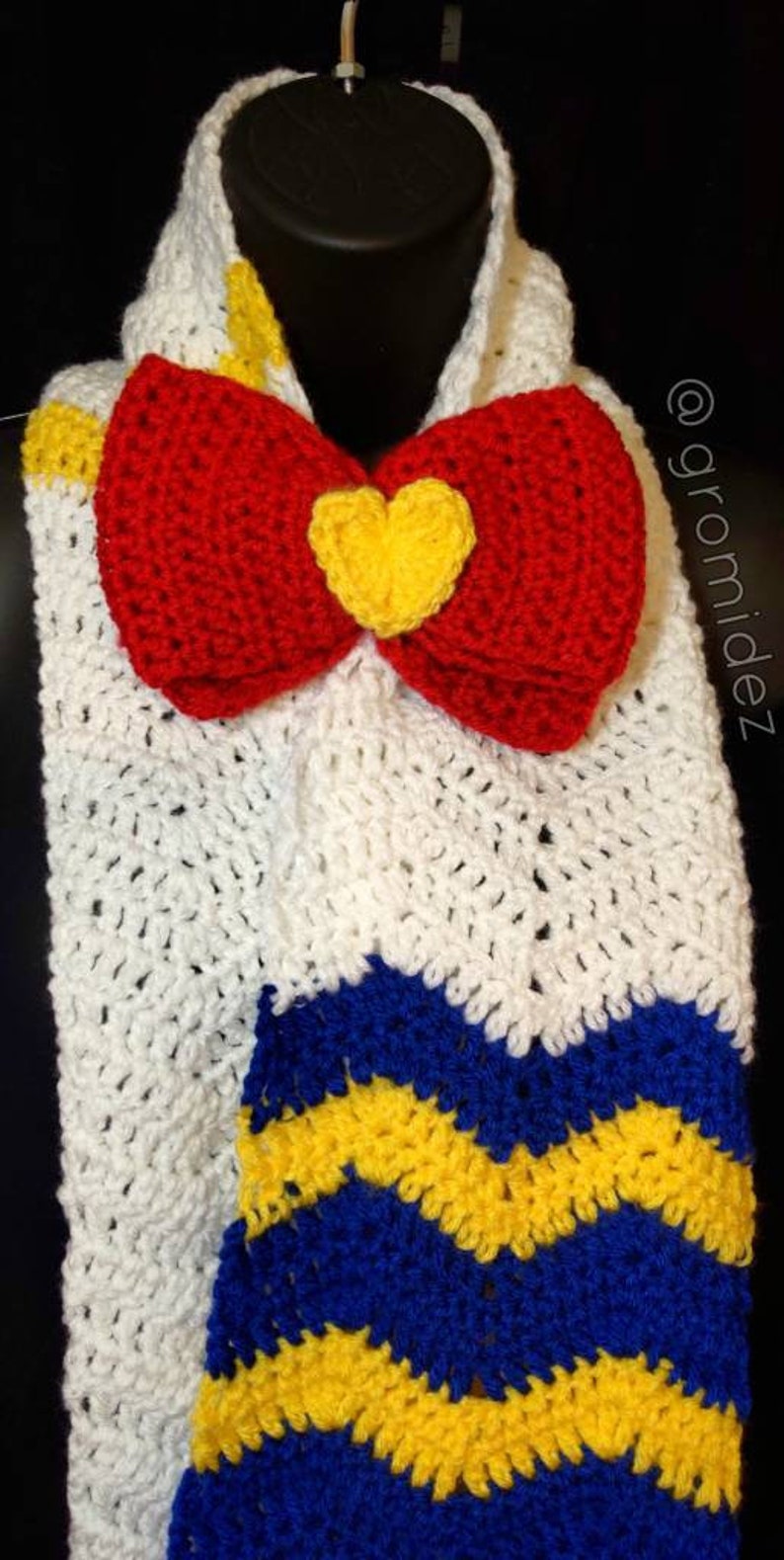 May include: A white crocheted scarf with a red bow and a yellow heart in the center. The scarf has blue and yellow stripes at the bottom.