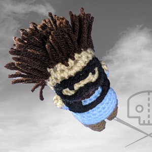 Puede incluir: Un juguete de peluche tejido a crochet con cabello marrón, una máscara negra y una camisa azul. El juguete está sobre un fondo de cielo nublado.