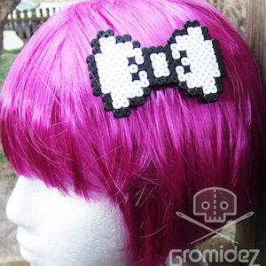 Puede incluir: Una pinza para el pelo con forma de lazo pixelado en blanco y negro. El lazo está hecho de pequeñas cuentas blancas con cuentas negras que delinean la forma.