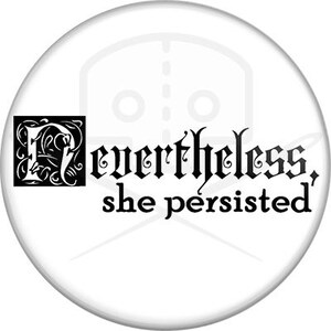 May include: A white button with black text that reads "Nevertheless, she persisted". The text is in a decorative font and includes a stylized letter "N" in a box.