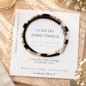 May include: A beaded bracelet featuring black, gold, and white beads arranged in a Morse code pattern. The bracelet is displayed on a white card with the text I CAN DO HARD THINGS, Ardent Fondness, and Morse Code Bracelet printed on it.