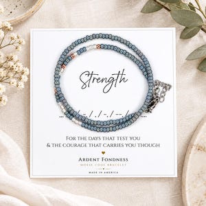 May include: A Morse code bracelet featuring blue, silver, and copper-colored beads on a white display card. The card displays the word Strength, Morse code dots and dashes, and the text For the days that test you & the courage that carries you though.