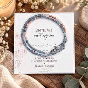 May include: A beaded bracelet featuring blue and metallic rose gold seed beads arranged in a circular pattern on a white display card. The card includes the text "UNTIL WE meet again," Morse code symbols, and "A QUIET REMINDER THAT LOVE NEVER LEAVES."