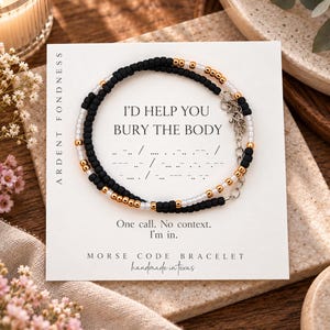 May include: A beaded bracelet featuring black, white, and metallic gold beads arranged in a pattern. The bracelet is displayed on a white card with the text I'D HELP YOU BURY THE BODY, Morse code symbols, and the phrase One call. No context. I'm in.