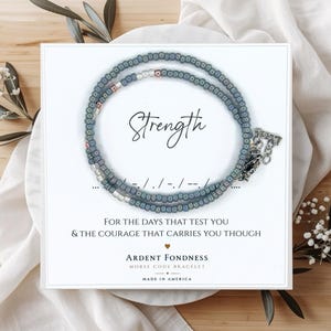 May include: A beaded bracelet featuring blue and metallic silver beads arranged in a Morse code pattern. The bracelet is displayed on a white card with the text Strength, For the days that test you & the courage that carries you though, Ardent Fondness, and Morse Code Bracelet.