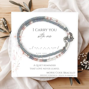 May include: A Morse code bracelet with a message, displayed on a white card with the text "I carry you with me." The bracelet is made of small, colorful beads. The card also includes the text "A quiet reminder that love never leaves."