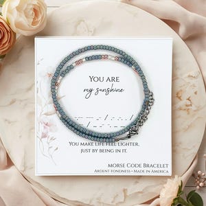 May include: A beaded Morse code bracelet in shades of blue, pink, and silver, displayed on a white card with the text "YOU ARE my sunshine". The card also includes the message "YOU MAKE LIFE FEEL LIGHTER, JUST BY BEING IN IT."