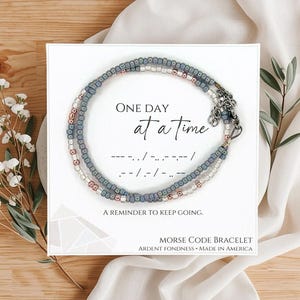 May include: A beaded Morse code bracelet with the message "One Day at a Time" on a white card. The bracelet features blue, white, and rose gold beads. The card also includes the Morse code translation and the phrase "A reminder to keep going."