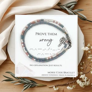 May include: A Morse code bracelet with a message, displayed on a white card. The bracelet is made of small, colorful beads in shades of blue, white, and pink. The card reads "PROVE THEM wrong" and "NO EXPLANATION. JUST RESULTS."