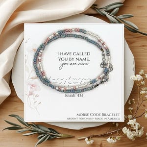 May include: A beaded Morse code bracelet with the text "I HAVE CALLED YOU BY NAME, you are mine" and "Isaiah 43:1". The bracelet is made of blue, white, and pink beads. The card reads "MORSE CODE BRACELET ARDENT FONDNESS MADE IN AMERICA".