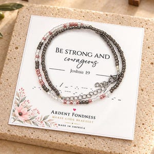 May include: A Morse code bracelet featuring metallic dark gray and light pink beads on a display card. The card reads BE STRONG AND courageous Joshua 1:9, Ardent Fondness Morse Code Bracelet, and Made in America, with a floral graphic at the bottom.