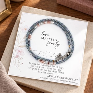 May include: A beaded bracelet featuring small blue, silver, and rose gold beads arranged in a Morse code pattern. The bracelet is displayed on a white card with the text "love MAKES US family" and a message about family, plus a Morse code sequence.